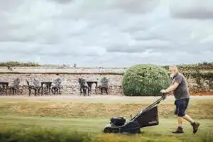 An older man with a gray beard pushes a lawn mower across a neatly trimmed grassy lawn in a garden, with a stone wall, patio tables and chairs, and a large round shrub under a cloudy sky.