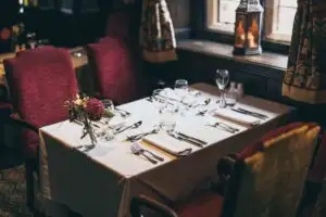Elegantly set dining table with white tablecloth, polished cutlery, wine glasses, and a floral centrepiece in a warmly lit restaurant.