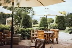 Outdoor garden terrace with wooden tables and chairs under large cream umbrellas, surrounded by manicured hedges and cone-shaped topiary trees in a landscaped countryside setting.