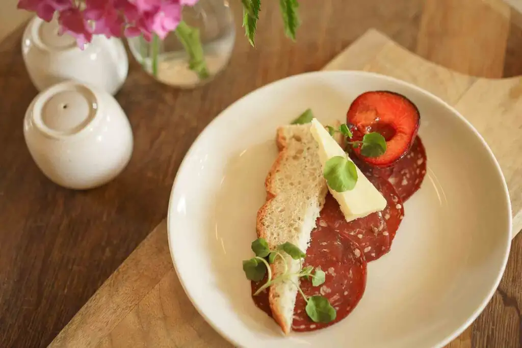 Slices of cured meat served with toasted bread, butter, microgreens, and a halved plum.