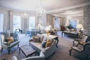 Spacious elegant lounge with chandeliers, upholstered seating, side tables with lamps, and large windows overlooking the gardens in a historic castle interior.