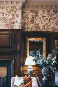 Detail of a traditional castle interior featuring ornate patterned wallpaper, dark wood panelling, a decorative fireplace, and a table lamp reflected in a gilt-framed mirror.
