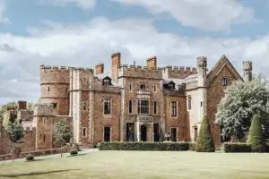 Large historic brick castle-style manor with round towers, tall chimneys, and decorative stone details, viewed from a wide lawn with trimmed hedges and trees under a partly cloudy sky.