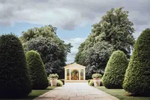 A tree-lined garden pathway leading to the classical outdoor ceremony pavilion at Rowton Castle, framed by manicured topiary and stone planters.