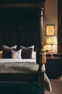 Traditional four-poster bed with carved wooden posts, layered decorative cushions, and a bedside table with a lit lamp in a classic castle bedroom.