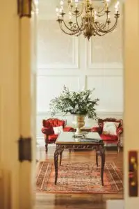 Elegant reception room with a chandelier, antique table topped with floral arrangement, red velvet sofas, and patterned rug, viewed through an open doorway.