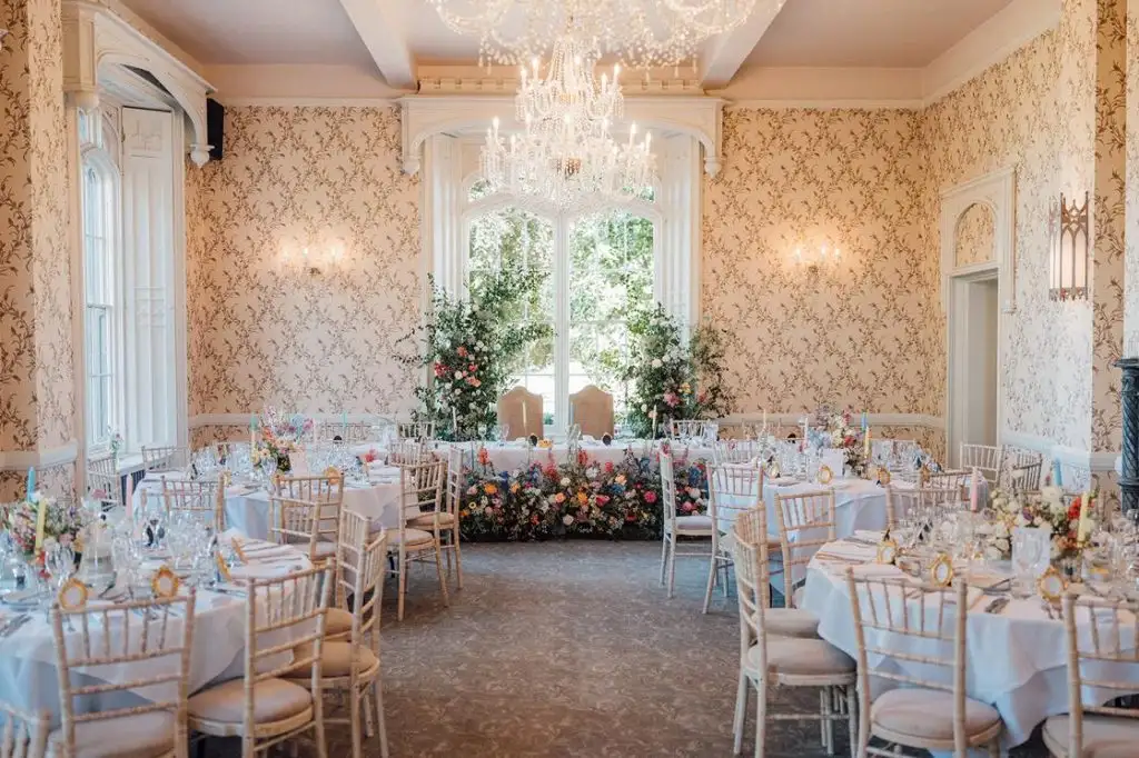 Elegant wedding reception room at Rowton Castle with round tables, Chiavari chairs, floral centrepieces, and a beautifully decorated top table beneath crystal chandeliers and tall windows.