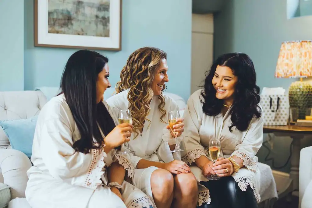 Three women in white robes enjoying champagne together in a relaxed bridal preparation setting