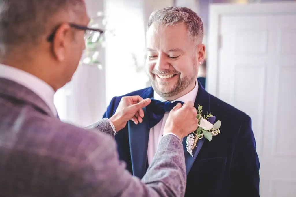 A groom smiling as his bow tie is adjusted before the ceremony