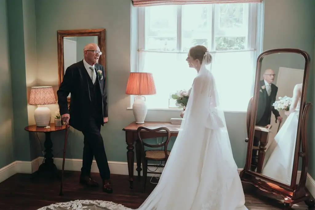 Bride in her wedding dress and veil standing by a window speaking with her father, who holds a walking cane, during a quiet pre-ceremony moment indoors.