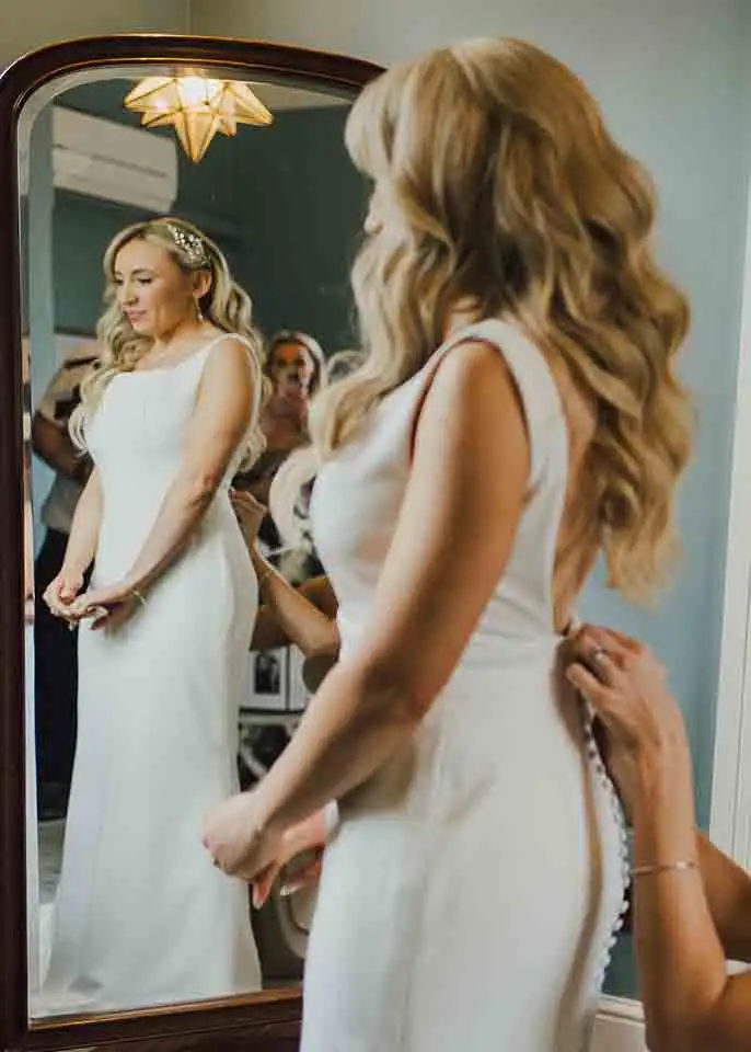 Bride standing in front of a mirror as someone fastens the back of her fitted wedding dress during morning preparations.