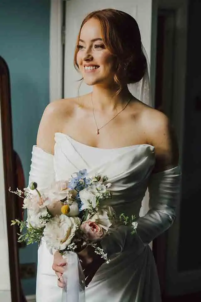 Bride in an elegant off-the-shoulder wedding dress holding a bouquet of pastel flowers while smiling softly near a window during wedding preparations.