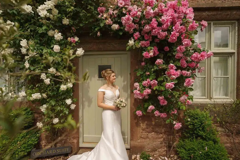 Bride in an off-the-shoulder wedding dress holding a bouquet while standing outside a cottage doorway framed by pink and white climbing roses.