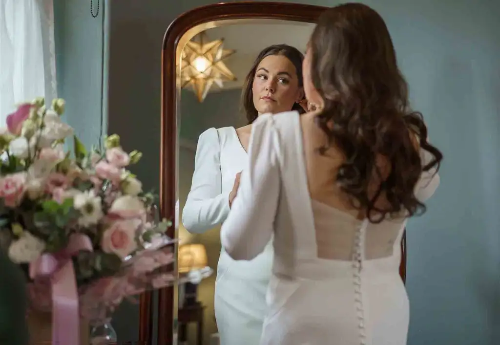 Bride in a long-sleeve wedding dress fastening her earrings while looking at her reflection in a mirror, with a bouquet of pink and white flowers nearby.