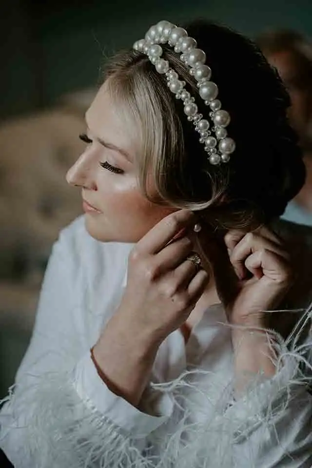 Bride wearing a pearl headband and robe adjusting her earrings while getting ready for her wedding day.
