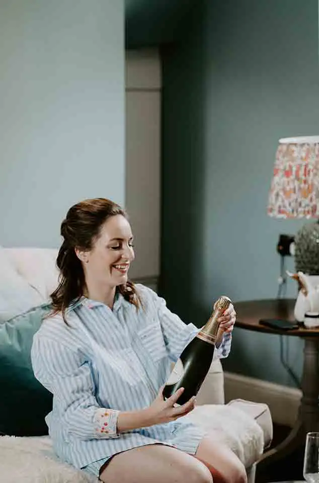 Bride in striped pajamas sitting on a sofa and smiling while opening a bottle of champagne during relaxed wedding morning preparations.