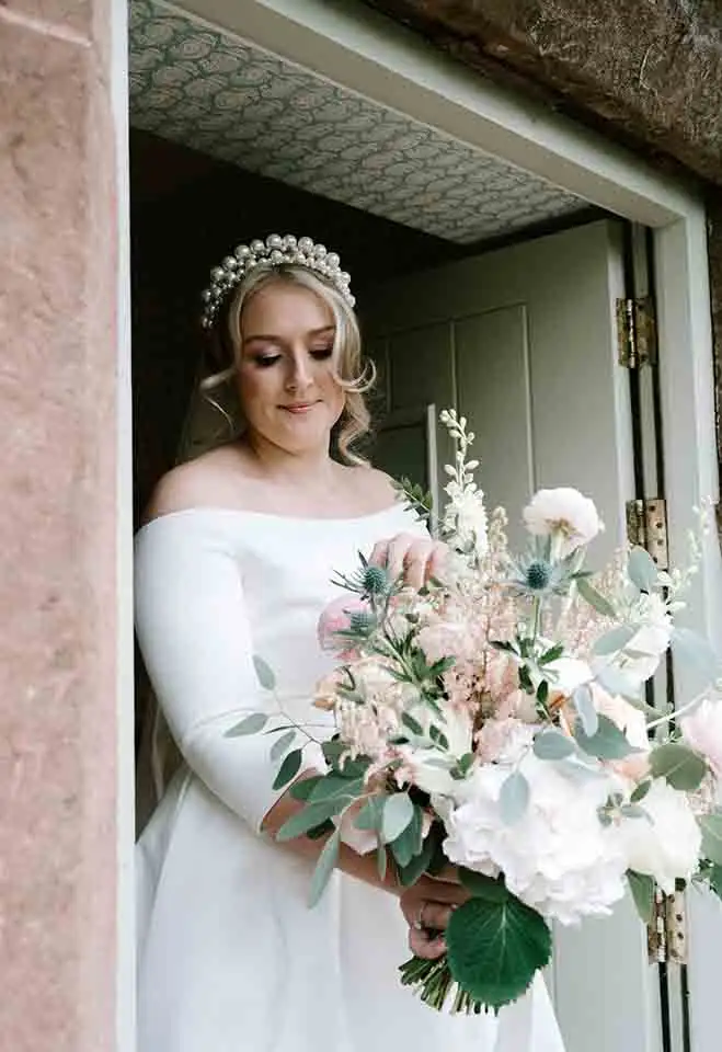 Bride in an off-the-shoulder wedding dress and pearl headband standing in a doorway while holding a pastel bouquet of roses and greenery.