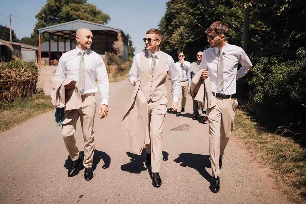 Group of groomsmen in light suits walking and chatting along a sunny country lane near Rowton Castle, surrounded by rural farm buildings and greenery.