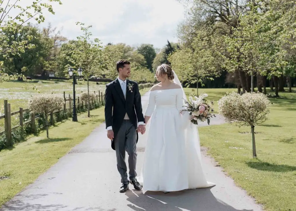 Bride and groom holding hands and walking down a tree-lined driveway at Rowton Castle, with spring blossom, green lawns, and countryside views in the background.