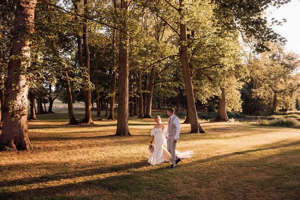 Bride and groom walking together across the lawns beneath tall trees at Rowton Castle, with warm golden sunlight filtering through the woodland.