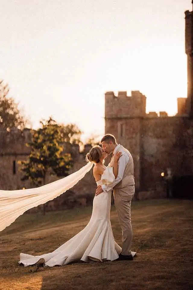 Bride and groom embracing at sunset in the grounds of Rowton Castle, with the bride’s long veil flowing behind her and the castle walls softly lit by golden evening light.