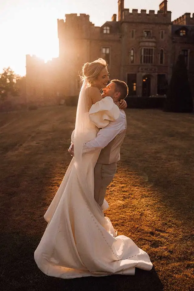 Groom lifting his bride in his arms in front of Rowton Castle at sunset, with warm golden light shining behind them and the bride’s veil softly glowing.