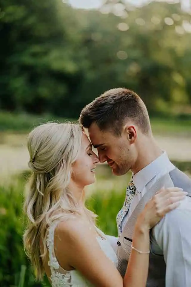 Close-up of a bride and groom embracing beside the lake at Rowton Castle, gently touching foreheads with soft greenery and water in the background.