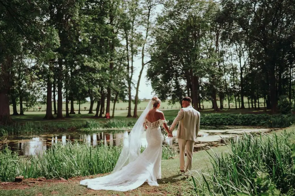Bride and groom holding hands and walking beside the lake in the grounds of Rowton Castle, with tall trees and landscaped gardens surrounding the water.