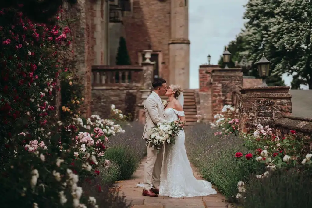 Bride and groom sharing a kiss on a garden pathway at Rowton Castle, surrounded by blooming roses and lavender with the historic castle building behind them.