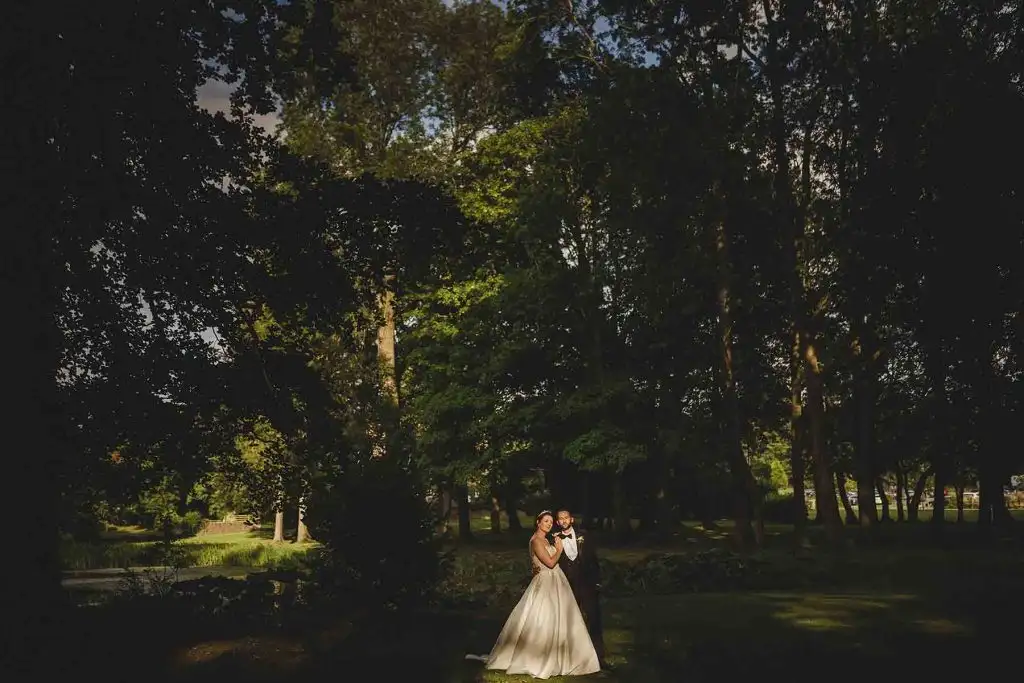 Bride and groom standing together beneath tall trees in the gardens at Rowton Castle, with warm evening light filtering through the woodland.