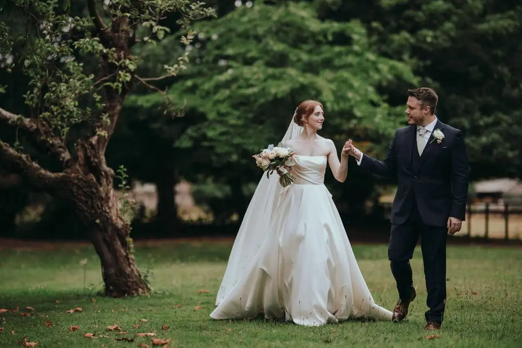 Bride holding a bouquet and groom in a dark suit walking hand in hand across the gardens at Rowton Castle with trees and greenery in the background.
