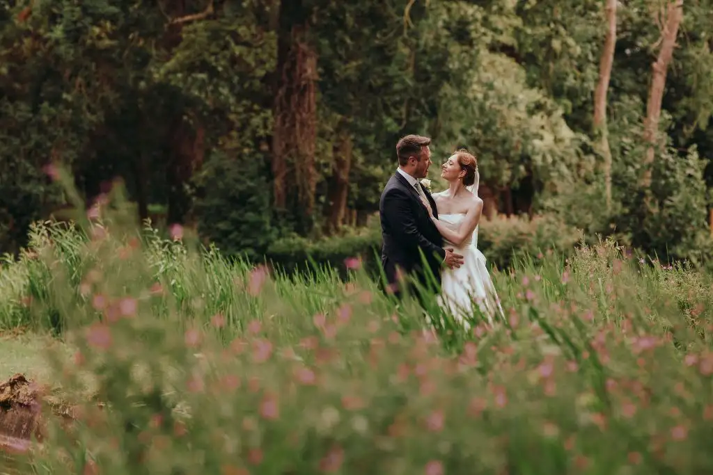 Bride and groom standing together in lush green gardens at Rowton Castle, surrounded by tall grasses and wildflowers with woodland trees in the background.