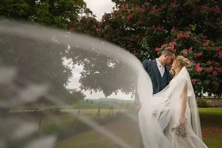Bride and groom sharing an intimate moment beneath a flowering tree at Rowton Castle, with the bride’s veil flowing softly across the foreground.