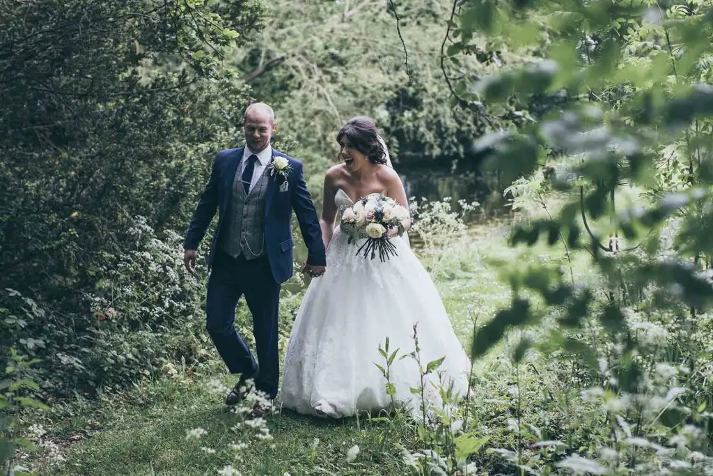 Bride holding a bouquet and groom in a blue suit walking hand in hand along a leafy lakeside path in the natural woodland grounds of Rowton Castle.