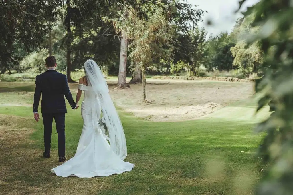 Bride and groom walking away hand in hand across the gardens at Rowton Castle, with the bride’s long veil flowing behind her and tall trees surrounding the grounds.