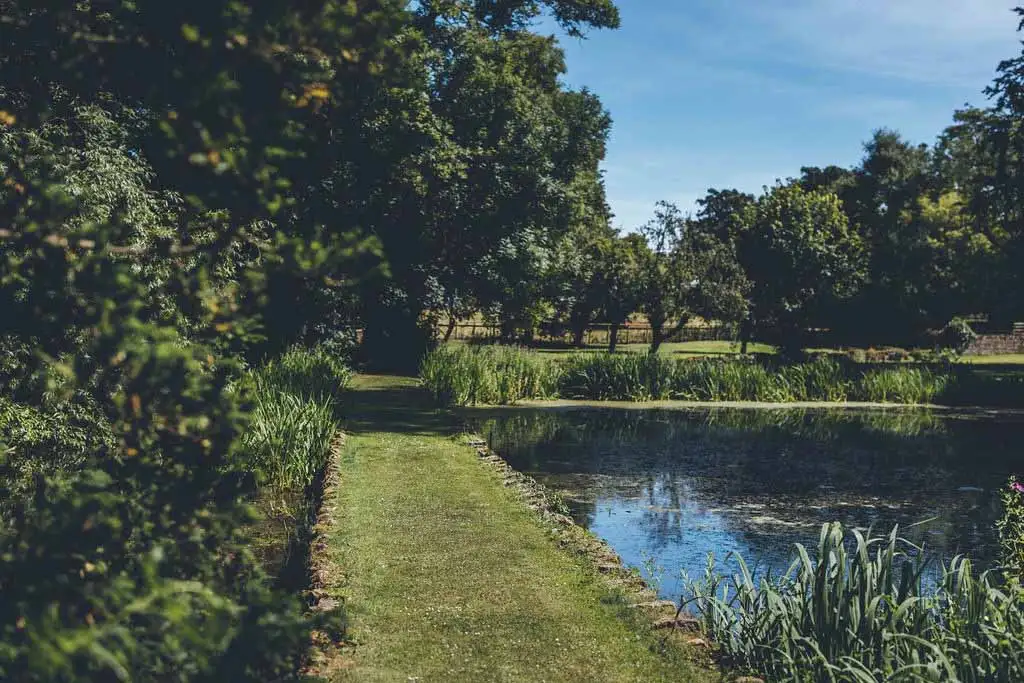 Scenic lakeside path bordered by lush greenery and trees in the landscaped gardens of Rowton Castle, with calm water reflecting the blue sky on a sunny day.