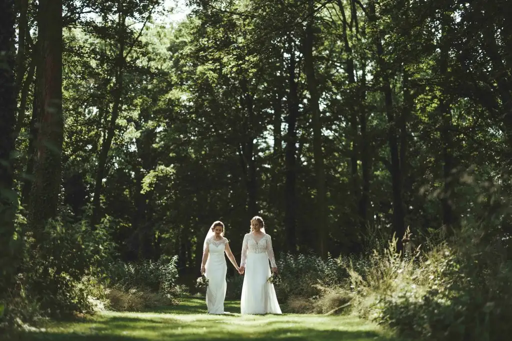 Two brides in wedding dresses walking hand in hand along a sunlit woodland path at Rowton Castle, surrounded by tall trees and soft green foliage.