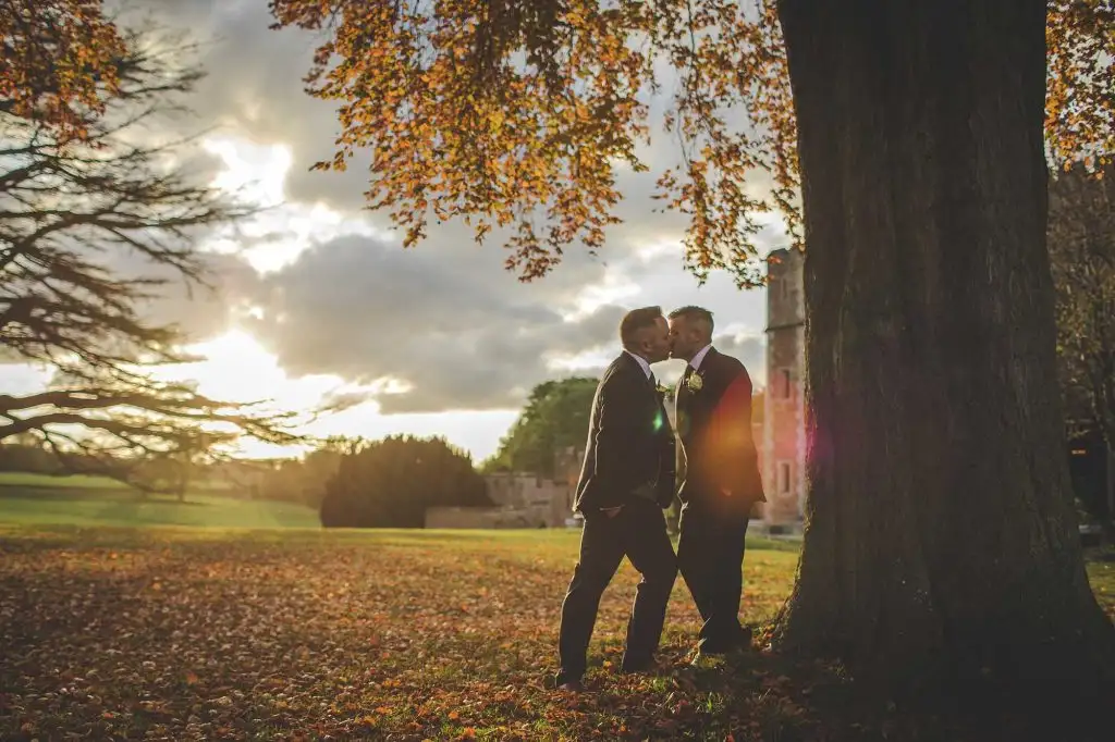 Two grooms sharing a kiss beneath a large tree at sunset at Rowton Castle, with golden light, autumn leaves on the ground, and the castle visible in the background.