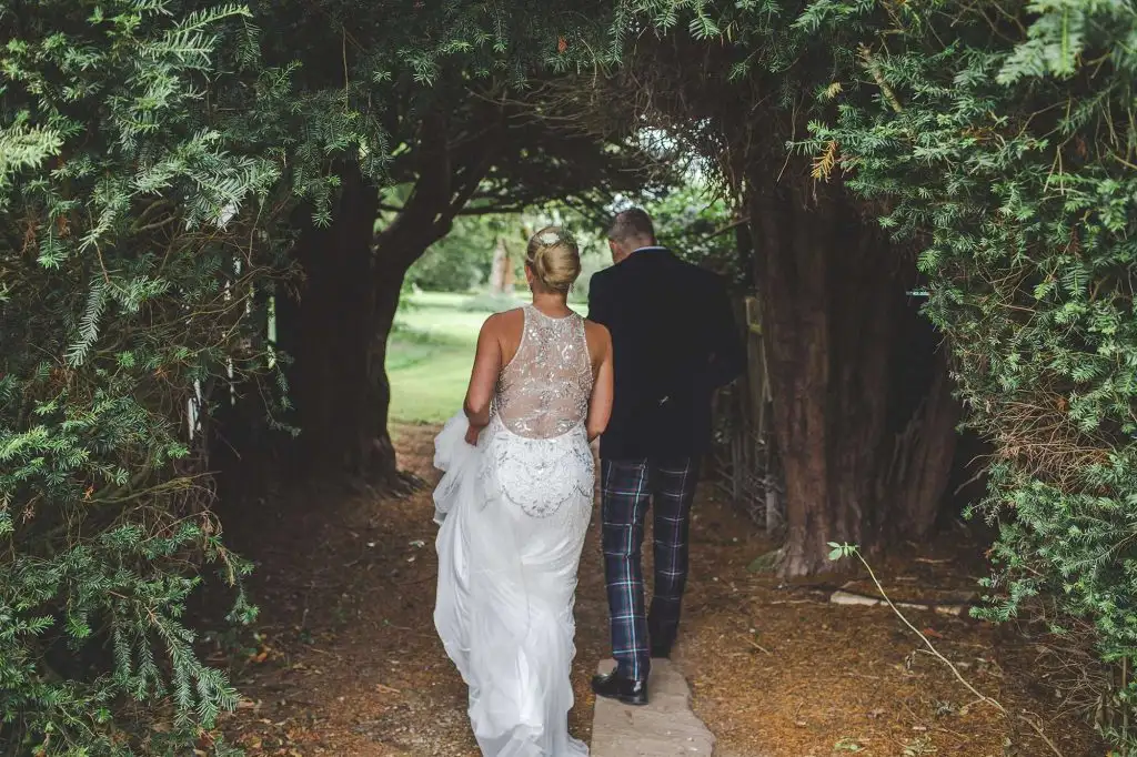 Bride in a lace-back wedding dress and groom in a kilt walking hand in hand along a shaded woodland path beneath arching trees at Rowton Castle grounds.