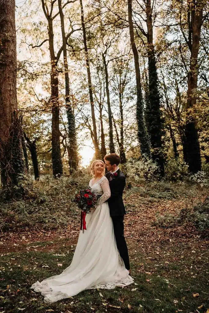 Bride and groom embracing in a sunlit woodland clearing at Rowton Castle, surrounded by tall trees and autumn leaves, with the bride holding a dark red bouquet.
