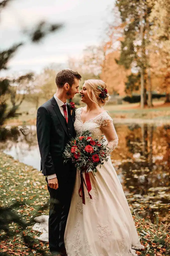 Bride and groom standing together beside the lake at Rowton Castle, surrounded by autumn trees, with the bride holding a red rose bouquet.
